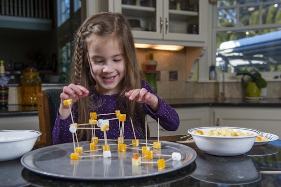 young girl with braid in hair building a structure with toothpicks, mashmallows, and cheese